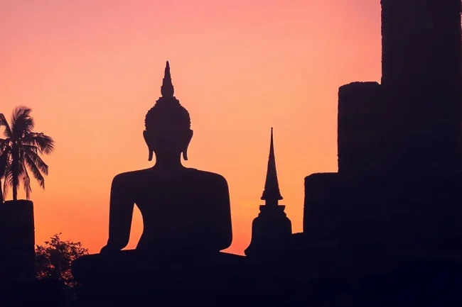 A Thai-style Buddha statue, seated, surrounded by poity stupas, silhouetted in front of a peach-colored sunset.