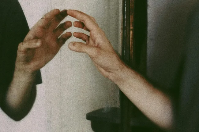A man's forearm and hand reach up to its reflection in a mirror. The image looks very like an oil painting.