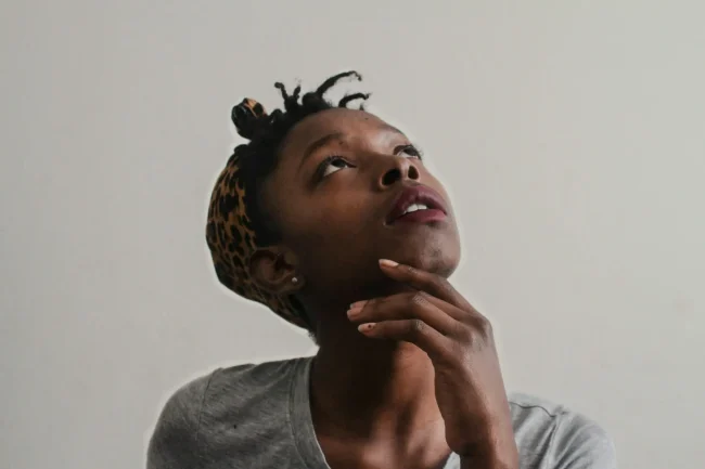 A black woman wearing a leopard print headscarf, under which mini braids are visible, has her hand on her chin and her head tilted back as she looks upward.